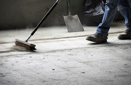 The Legs And Feet Of A Construction Worker Sweeping Up On Rough Concrete At A Job Site Using A Large Broom